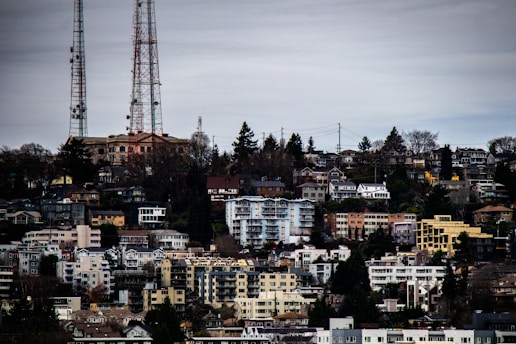 A dense urban neighborhood with various multi-story residential buildings and houses on a hillside. Distant radio towers rise above the treeline in the background. The scene is overcast, and the architecture features diverse styles and colors, adding to the complexity of the landscape.