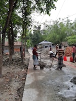 Several men are working on a construction site in a rural area, mixing cement or concrete on a paved pathway. The scene is surrounded by greenery, with trees lining the pathway. Piles of bricks and construction materials are visible, and small houses with tin roofs are in the background.