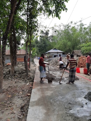 Several men are working on a construction site in a rural area, mixing cement or concrete on a paved pathway. The scene is surrounded by greenery, with trees lining the pathway. Piles of bricks and construction materials are visible, and small houses with tin roofs are in the background.