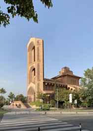 A large, impressive brick church with a tall bell tower featuring a cross at the top. The architecture showcases arches and intricate brickwork. Surrounding the church are trees and greenery, with a clear, blue sky overhead. A crosswalk and road signs are visible in the foreground.
