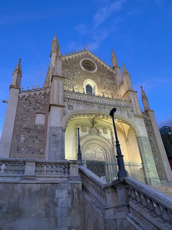 The image features a historic Gothic-style church facade, characterized by intricate stonework and pointed spires. The entrance is grand, with detailed carvings above the doorway. The structure is illuminated, highlighting its architectural details. The sky is clear with a soft blue hue, suggesting early evening or morning.