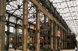 A large industrial-style building features exposed metal beams and columns, with a glass roof allowing natural light to illuminate the interior. The structure includes wooden elements with modern touches, such as glass walls and greenery. People can be seen walking around the area, and colorful banners hang from the upper parts of the building.