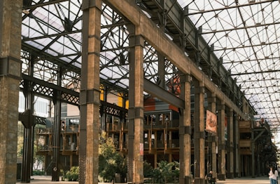 A large industrial-style building features exposed metal beams and columns, with a glass roof allowing natural light to illuminate the interior. The structure includes wooden elements with modern touches, such as glass walls and greenery. People can be seen walking around the area, and colorful banners hang from the upper parts of the building.