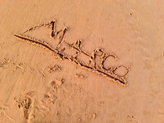 The image depicts the word 'Marco' written in the sand, with visible grains and textures. The surrounding area is smooth, showing gentle curves and uneven imprints in the sand.