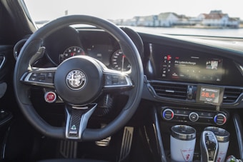 A view of a car's interior highlighting the steering wheel with the Alfa Romeo logo, dashboard displays, and various control features. The gear shift and cup holders are prominently visible, alongside digital displays showing information such as temperature and radio stations.