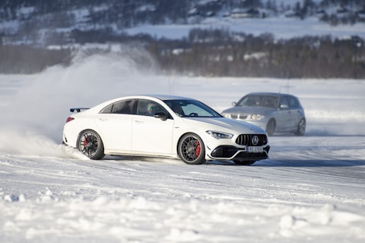 A white car is drifting on a snowy surface, creating a cloud of snow behind it. Another car, slightly blurred, follows in the background. The landscape features snow-covered fields and distant mountains.