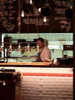 A chef wearing a striped apron is working behind a wooden counter in a cozy restaurant kitchen. The menu is written on black chalkboards above the counter with various dishes and prices. The lighting is warm and soft, creating an inviting atmosphere. Hanging lamps illuminate the workspace, while stacks of plates and kitchen equipment are visible in the background.