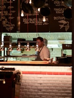 A chef wearing a striped apron is working behind a wooden counter in a cozy restaurant kitchen. The menu is written on black chalkboards above the counter with various dishes and prices. The lighting is warm and soft, creating an inviting atmosphere. Hanging lamps illuminate the workspace, while stacks of plates and kitchen equipment are visible in the background.
