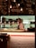 A chef wearing a striped apron is working behind a wooden counter in a cozy restaurant kitchen. The menu is written on black chalkboards above the counter with various dishes and prices. The lighting is warm and soft, creating an inviting atmosphere. Hanging lamps illuminate the workspace, while stacks of plates and kitchen equipment are visible in the background.