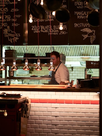 A chef wearing a striped apron is working behind a wooden counter in a cozy restaurant kitchen. The menu is written on black chalkboards above the counter with various dishes and prices. The lighting is warm and soft, creating an inviting atmosphere. Hanging lamps illuminate the workspace, while stacks of plates and kitchen equipment are visible in the background.