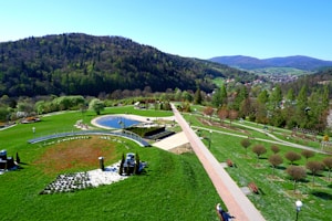A scenic park landscape with lush green grass, neatly arranged walking paths, and a decorative circular pond. The park is surrounded by hills covered in dense forest, with a small town visible in the background. Trees with full foliage line the paths, and there are benches and streetlights placed throughout the area.
