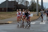 A group of runners participating in a road race, with some athletes wearing red and white sports outfits. The scene takes place on a wet road near a crosswalk, with trees and a house visible in the background. The sky is overcast, suggesting cloudy weather.