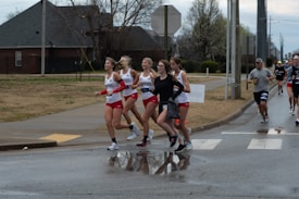 A group of runners participating in a road race, with some athletes wearing red and white sports outfits. The scene takes place on a wet road near a crosswalk, with trees and a house visible in the background. The sky is overcast, suggesting cloudy weather.