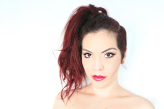 A person with striking makeup and bold red lipstick gazes directly at the camera. The hair is styled in a voluminous updo with rich, dark red tones.
