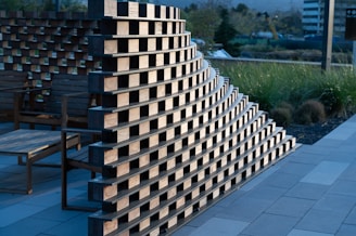 A curved wooden structure built from a pattern of stacked, rectangular blocks on a paved outdoor area. Wooden chairs are placed nearby, and there is grass and landscaping in the background, with buildings visible in the distance.