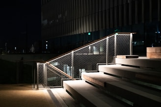 Concrete steps with wooden trim are illuminated by artificial lighting at night. The area is bordered by a metal railing with mesh wire. The building facade in the background is dark, allowing the lit railing and steps to stand out.
