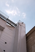 A construction site with scaffolding and protective white sheeting covering part of a building, set against a blue sky with scattered clouds.
