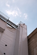 A construction site with scaffolding and protective white sheeting covering part of a building, set against a blue sky with scattered clouds.