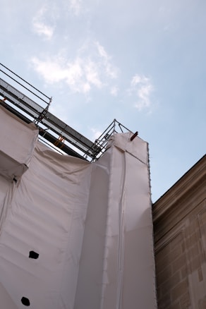 A construction site with scaffolding and protective white sheeting covering part of a building, set against a blue sky with scattered clouds.