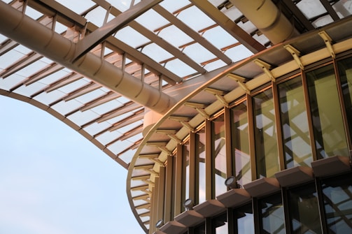 Detailed view of a modern architectural structure featuring a combination of glass and metal. Curved beams and large glass windows dominate the scene, with light reflections creating intricate patterns on the surface.