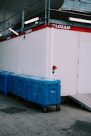 A row of blue industrial waste bins is aligned against a white portable building with the 'LOXAM' logo displayed on the side. Above the bins are various metal structures and lighting fixtures. The ground is paved with asphalt, and there's a ramp leading to a doorway on the building.
