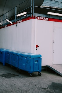 A row of blue industrial waste bins is aligned against a white portable building with the 'LOXAM' logo displayed on the side. Above the bins are various metal structures and lighting fixtures. The ground is paved with asphalt, and there's a ramp leading to a doorway on the building.