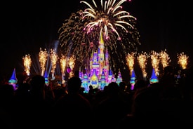 A brightly lit castle serves as the centerpiece, illuminated by vibrant lighting that creates a magical appearance. Surrounding the castle, fireworks explode in the night sky, lighting up the area with golden sparks. Silhouetted figures of people watching can be seen in the foreground, adding to the sense of wonder and celebration.
