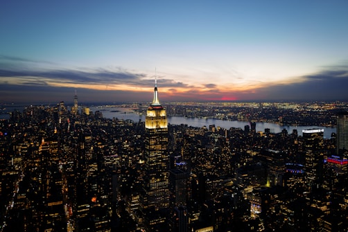A panoramic view of a bustling cityscape during sunset, with a prominent skyscraper illuminated in the foreground. The skyline is filled with numerous buildings, all twinkling with lights. The sky displays a gradient of blue, yellow, and hints of red from the setting sun.