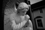 A monochrome statue of an angel kneeling in a prayerful pose with folded hands. The figure has large, detailed wings and long, wavy hair, and is positioned outdoors near a building.