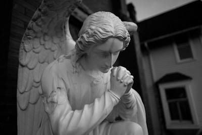 A monochrome statue of an angel kneeling in a prayerful pose with folded hands. The figure has large, detailed wings and long, wavy hair, and is positioned outdoors near a building.