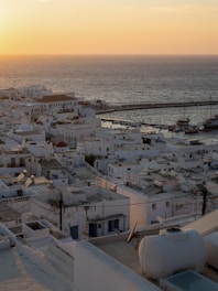 A scenic view of a coastal town with white-washed buildings packed closely together. The sea stretches into the horizon as the sun sets, casting a warm, golden glow over the landscape. Boats are docked at the harbor in the distance, and the architecture is typical of Cycladic style.