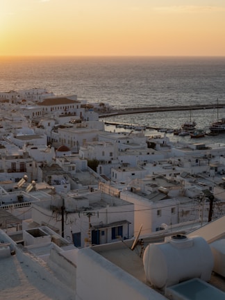 A scenic view of a coastal town with white-washed buildings packed closely together. The sea stretches into the horizon as the sun sets, casting a warm, golden glow over the landscape. Boats are docked at the harbor in the distance, and the architecture is typical of Cycladic style.