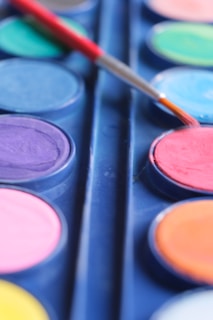 A close-up view of a watercolor paint set with a paintbrush resting on one of the vibrant, circular pans of color. The palette is filled with a variety of hues including blue, green, purple, pink, orange, and yellow.