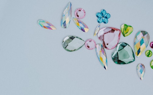 A collection of assorted, colorful gemstones in various shapes and sizes is laid out on a light gray background. The gems include teardrops, hearts, and flower-like shapes. They vary in color, featuring shades of pink, green, blue, and multicolored facets.