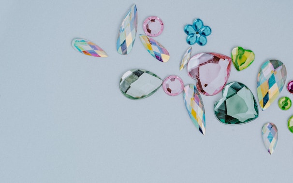 A collection of assorted, colorful gemstones in various shapes and sizes is laid out on a light gray background. The gems include teardrops, hearts, and flower-like shapes. They vary in color, featuring shades of pink, green, blue, and multicolored facets.