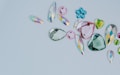 A collection of assorted, colorful gemstones in various shapes and sizes is laid out on a light gray background. The gems include teardrops, hearts, and flower-like shapes. They vary in color, featuring shades of pink, green, blue, and multicolored facets.