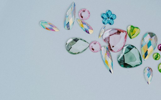 A collection of assorted, colorful gemstones in various shapes and sizes is laid out on a light gray background. The gems include teardrops, hearts, and flower-like shapes. They vary in color, featuring shades of pink, green, blue, and multicolored facets.