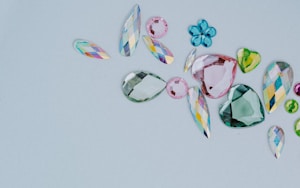 A collection of assorted, colorful gemstones in various shapes and sizes is laid out on a light gray background. The gems include teardrops, hearts, and flower-like shapes. They vary in color, featuring shades of pink, green, blue, and multicolored facets.