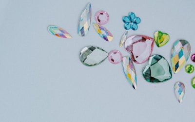 A collection of assorted, colorful gemstones in various shapes and sizes is laid out on a light gray background. The gems include teardrops, hearts, and flower-like shapes. They vary in color, featuring shades of pink, green, blue, and multicolored facets.