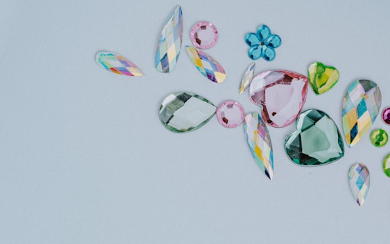A collection of assorted, colorful gemstones in various shapes and sizes is laid out on a light gray background. The gems include teardrops, hearts, and flower-like shapes. They vary in color, featuring shades of pink, green, blue, and multicolored facets.