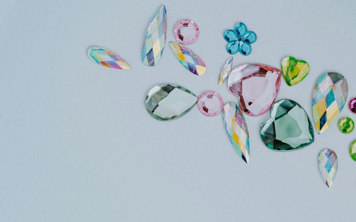 A collection of assorted, colorful gemstones in various shapes and sizes is laid out on a light gray background. The gems include teardrops, hearts, and flower-like shapes. They vary in color, featuring shades of pink, green, blue, and multicolored facets.