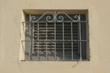 A small window with closed wooden shutters is covered by a decorative, slightly rusted metal grate featuring intricate scrollwork at the top. The window and grate are set into a light beige-colored wall with a rough, textured surface.