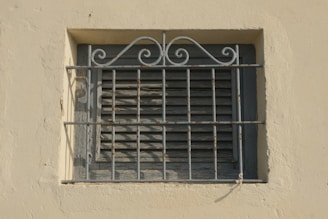 A small window with closed wooden shutters is covered by a decorative, slightly rusted metal grate featuring intricate scrollwork at the top. The window and grate are set into a light beige-colored wall with a rough, textured surface.