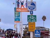 A variety of signs and a billboard are placed on a pole. The signs include a tsunami evacuation route, bike route, share the road, speed limit 5, and entrance signs. A colorful billboard advertises a local initiative called 'Santa Monica Shines' with the message 'Your Local Love, Eat, Shop, Stay.' Below the signs, there is a crowd of people walking, and in the background, there are some palm trees and a building. The sky is overcast.