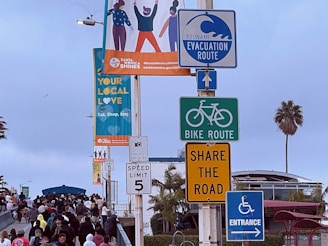 A variety of signs and a billboard are placed on a pole. The signs include a tsunami evacuation route, bike route, share the road, speed limit 5, and entrance signs. A colorful billboard advertises a local initiative called 'Santa Monica Shines' with the message 'Your Local Love, Eat, Shop, Stay.' Below the signs, there is a crowd of people walking, and in the background, there are some palm trees and a building. The sky is overcast.