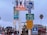 A variety of signs and a billboard are placed on a pole. The signs include a tsunami evacuation route, bike route, share the road, speed limit 5, and entrance signs. A colorful billboard advertises a local initiative called 'Santa Monica Shines' with the message 'Your Local Love, Eat, Shop, Stay.' Below the signs, there is a crowd of people walking, and in the background, there are some palm trees and a building. The sky is overcast.