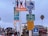 A variety of signs and a billboard are placed on a pole. The signs include a tsunami evacuation route, bike route, share the road, speed limit 5, and entrance signs. A colorful billboard advertises a local initiative called 'Santa Monica Shines' with the message 'Your Local Love, Eat, Shop, Stay.' Below the signs, there is a crowd of people walking, and in the background, there are some palm trees and a building. The sky is overcast.
