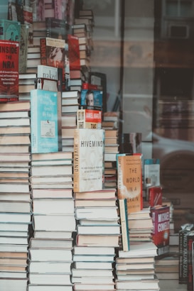 A large, towering stack of books displayed prominently in a shop window. Various titles and covers are visible, including Inside the NRA and The World in a Grain. The books are arranged haphazardly, creating a visually striking display. The reflection of the street outside can be seen in the window glass.