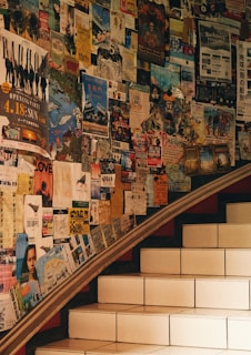A set of stairs bordered by a wall covered in numerous colorful posters and flyers, creating a collage effect. The posters include a variety of information such as events, concerts, and announcements. The ambiance is warm due to the earthy lighting.