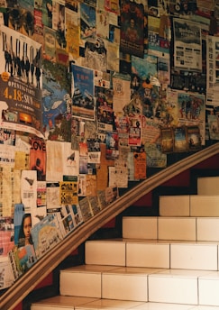 A set of stairs bordered by a wall covered in numerous colorful posters and flyers, creating a collage effect. The posters include a variety of information such as events, concerts, and announcements. The ambiance is warm due to the earthy lighting.